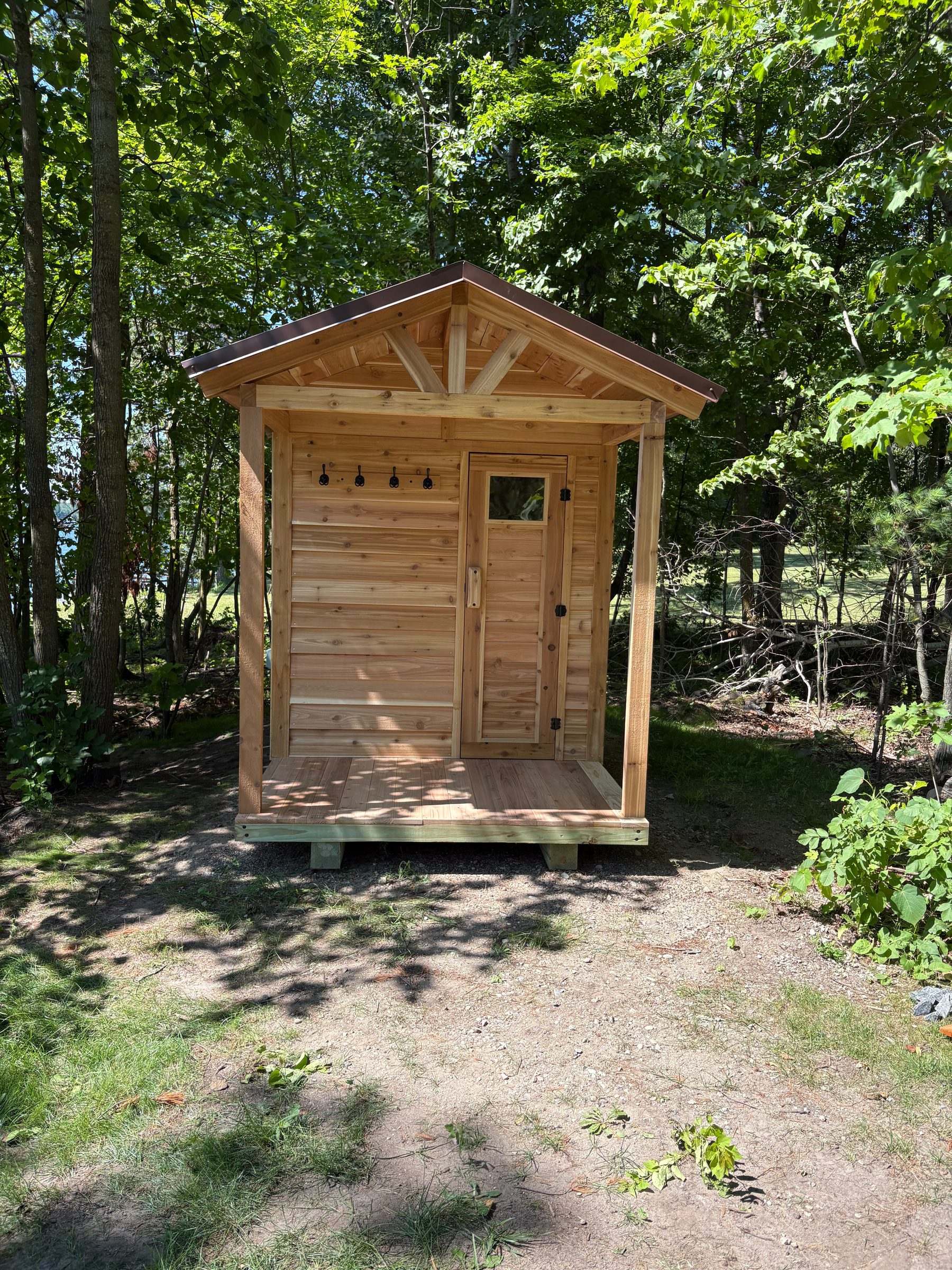 Front view of a cedar outdoor sauna with a covered front porch and towel hooks, set in the woods