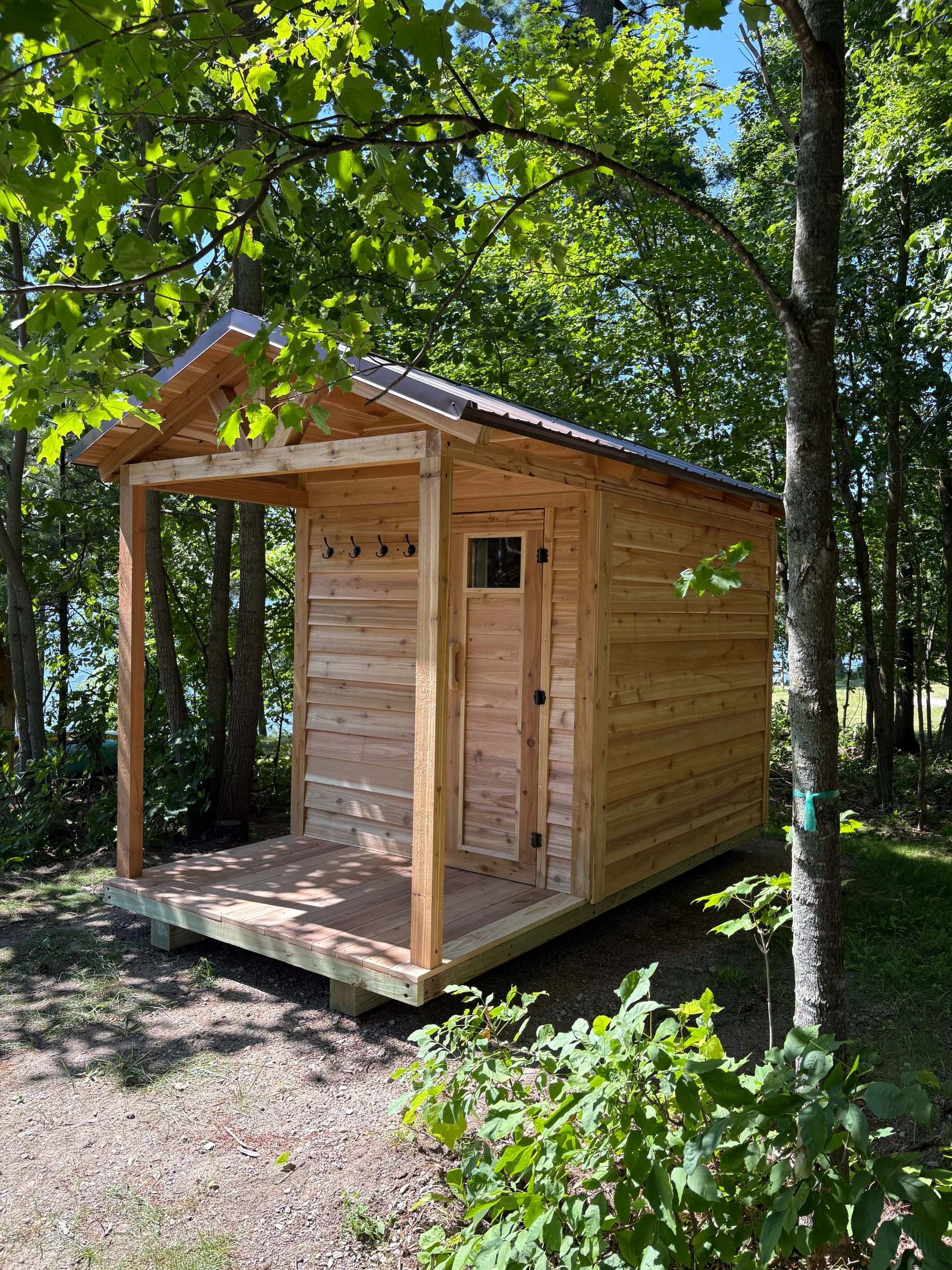Cedar outdoor sauna with a covered porch under trees on a lake lot