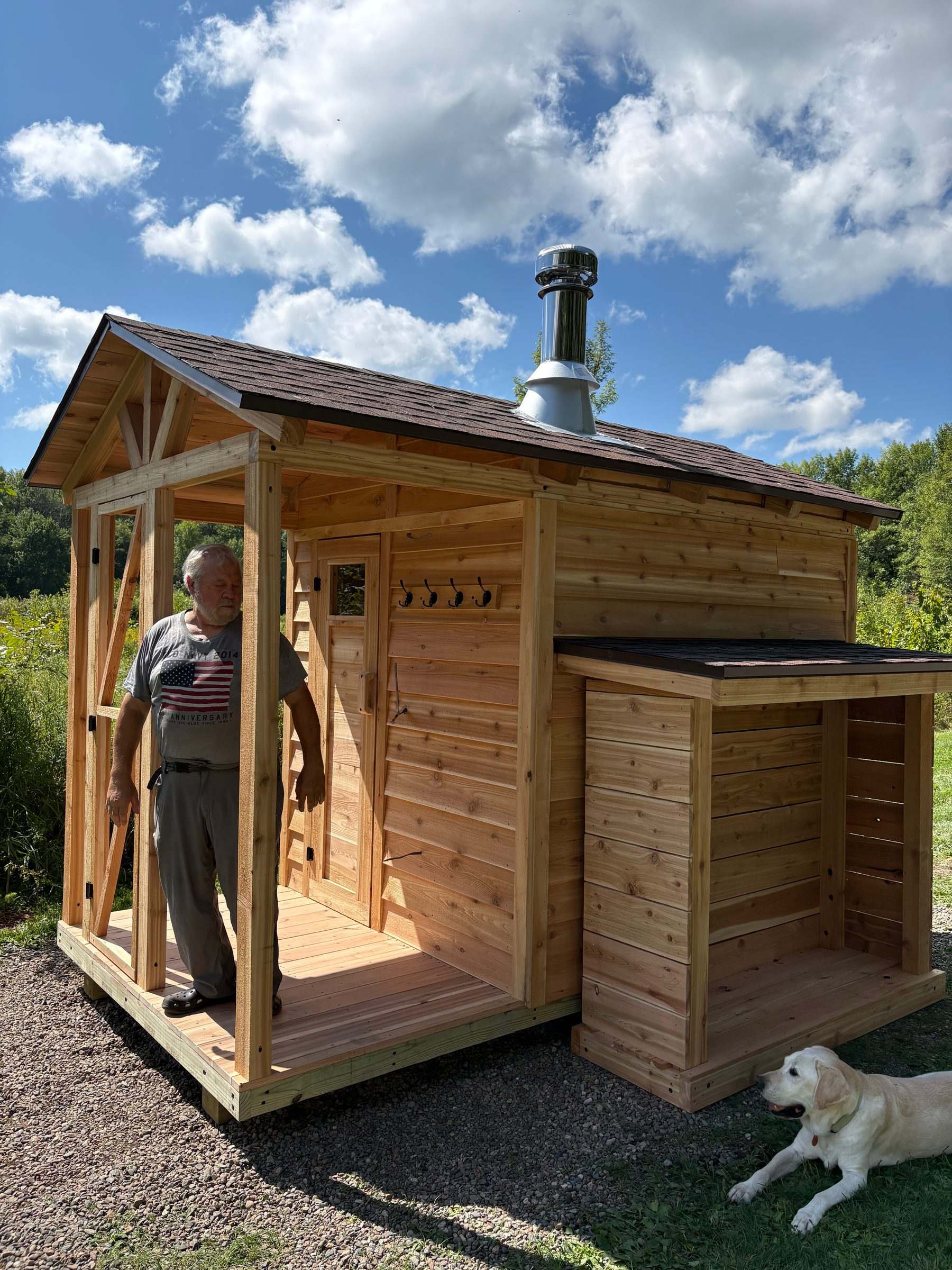 Cedar outdoor sauna with an attached woodshed, shingle roof and stainless chimney, with a man on the porch and a dog in the grass