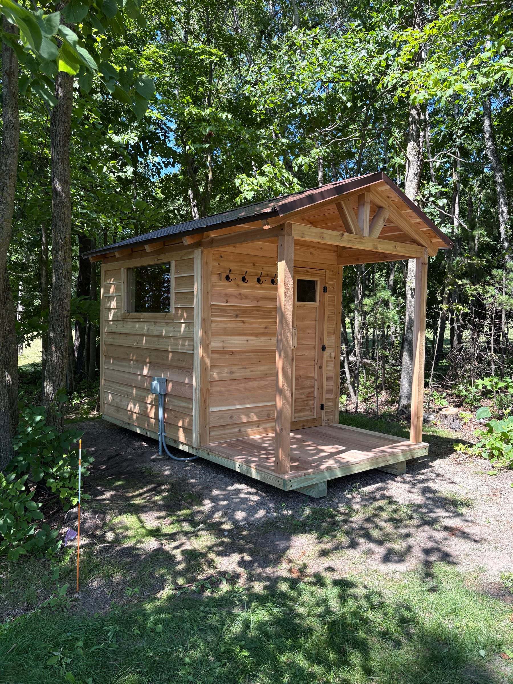 Cedar outdoor sauna with a wide side window, covered porch and an electrical disconnect for its electric heater