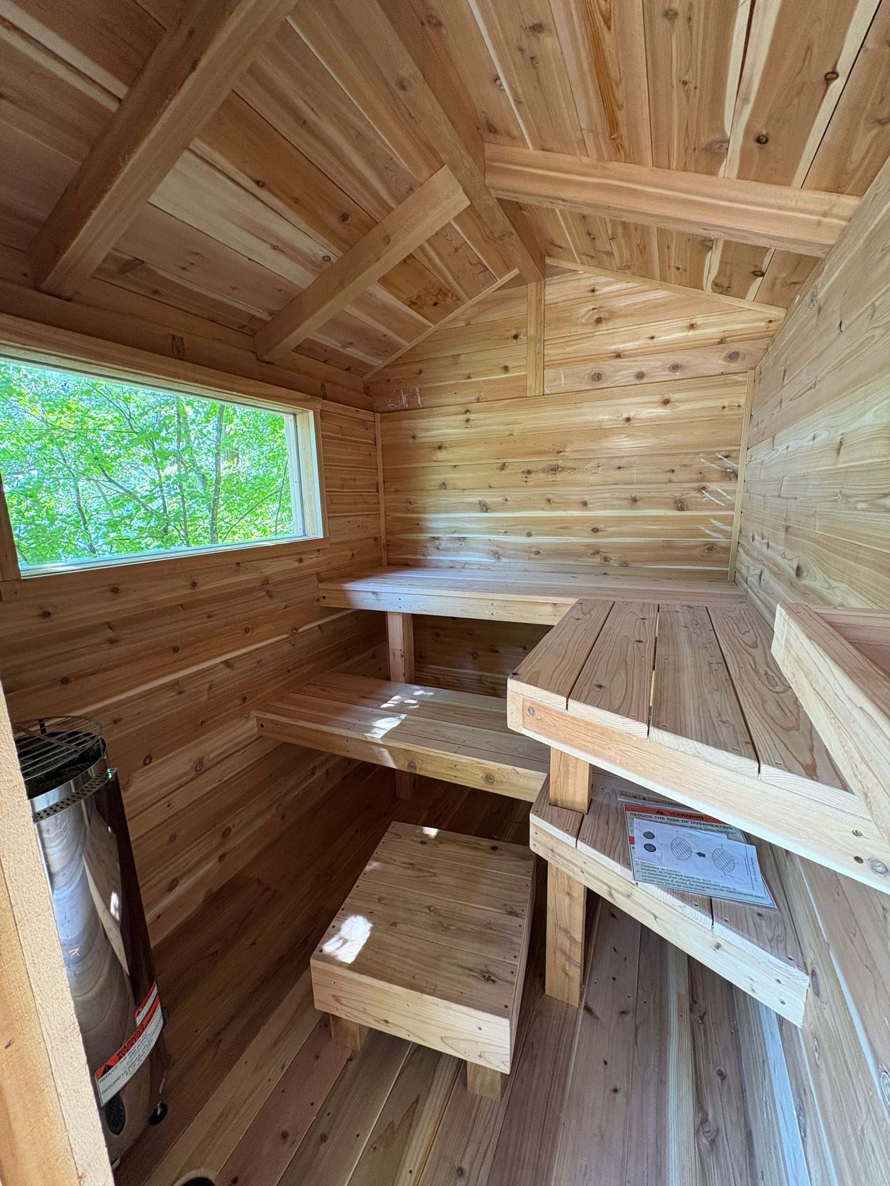 Cylindrical electric sauna heater beside tiered cedar benches and a window in a small outdoor sauna
