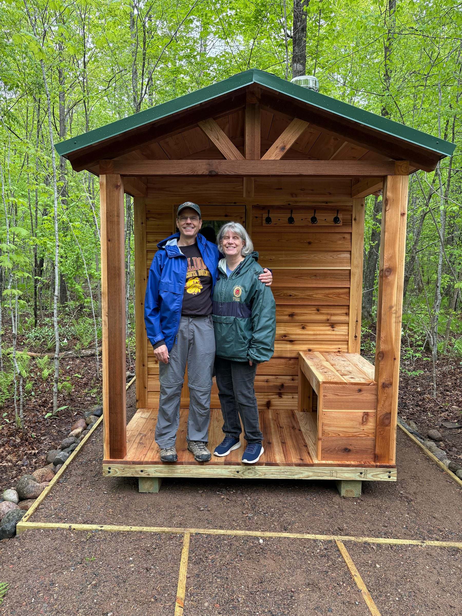 Two customers standing on the open covered porch of their new cedar sauna, with a cedar bench and towel hooks