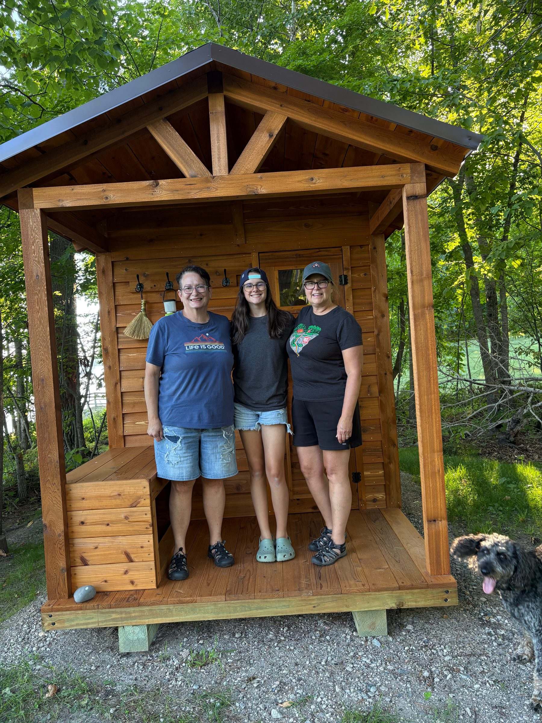Three smiling customers and their dog on the porch of their stained cedar sauna