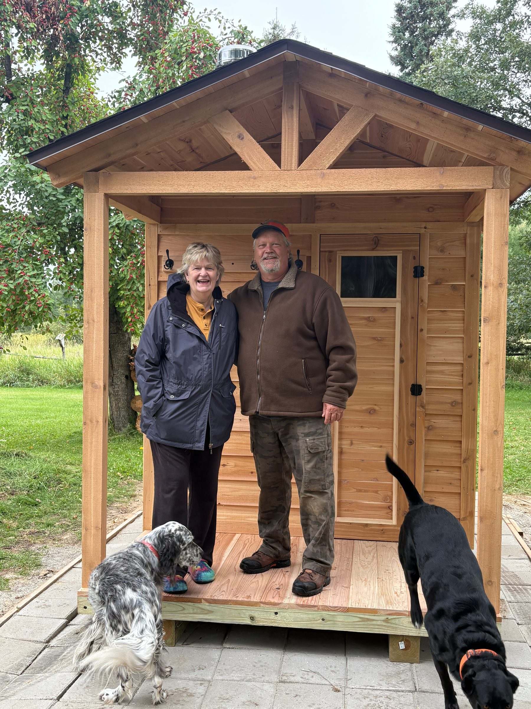 Two smiling customers and their dogs on the porch of their new cedar sauna, set on a paver pad