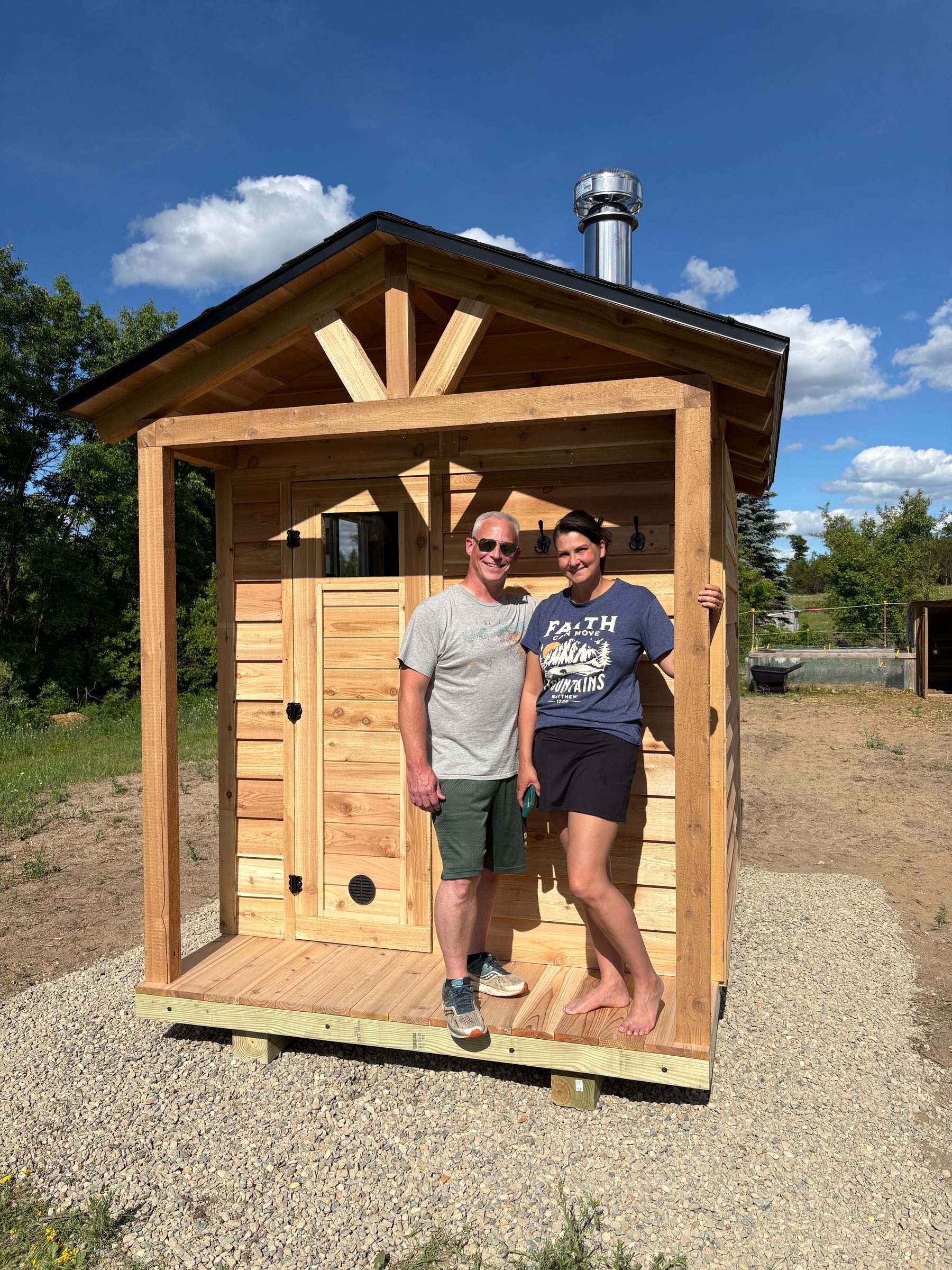 Two customers on the porch of their new wood-fired cedar sauna with a stainless chimney under a blue sky