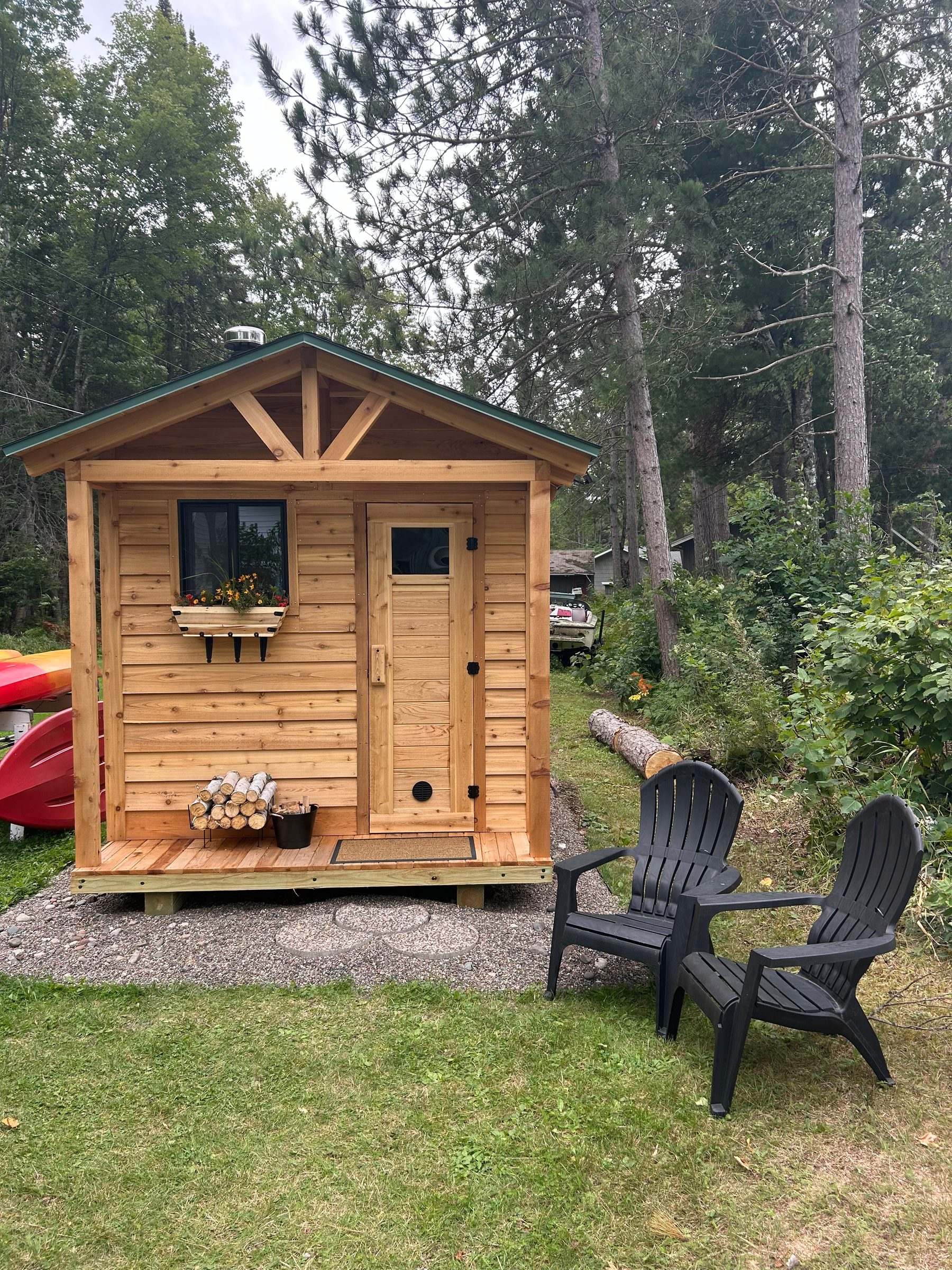 Cedar outdoor sauna with a small front porch, window flower box and stacked firewood at a lake cabin