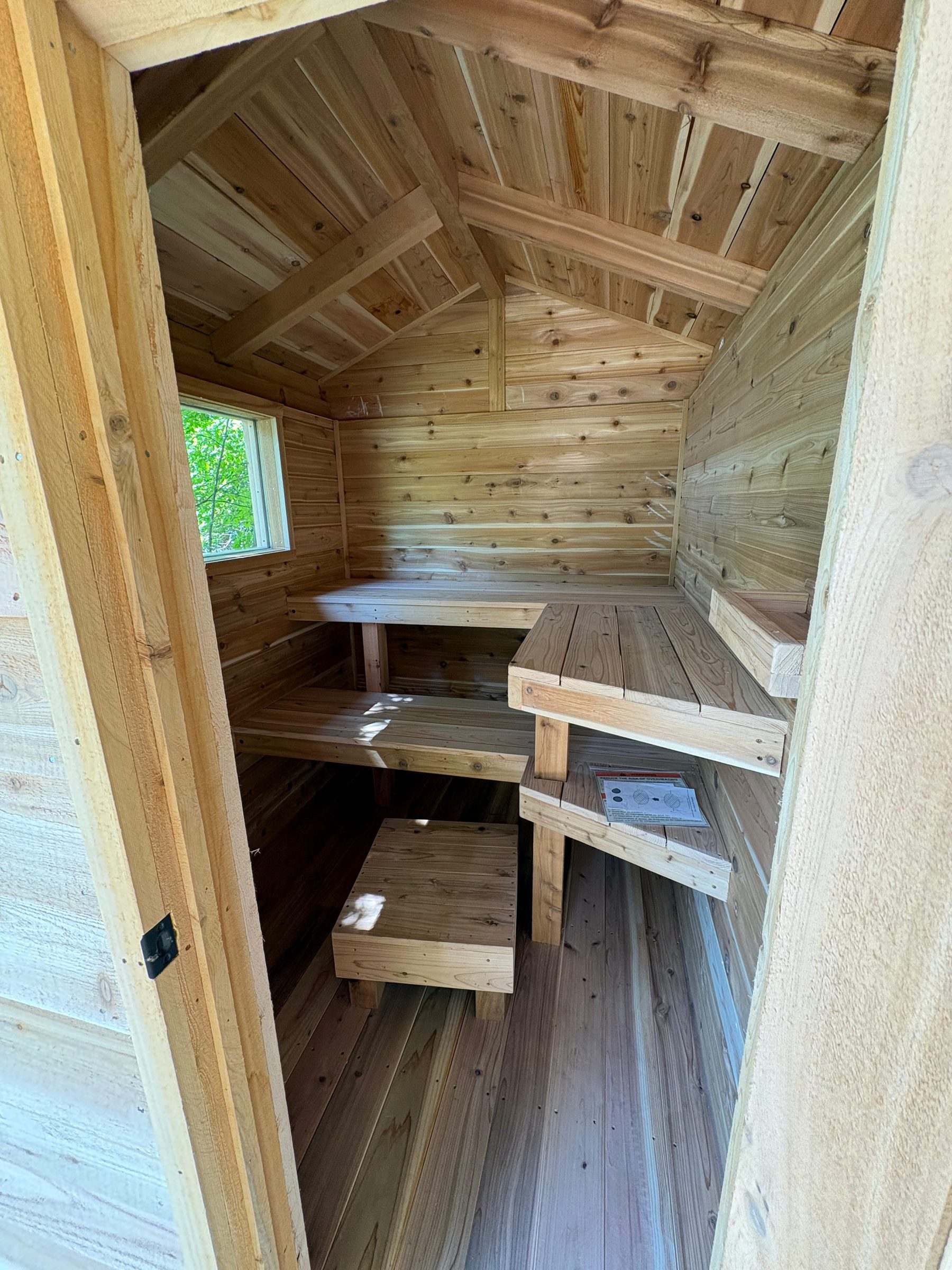 Inside a small cedar sauna: L-shaped upper and lower benches, a foot stool and a window