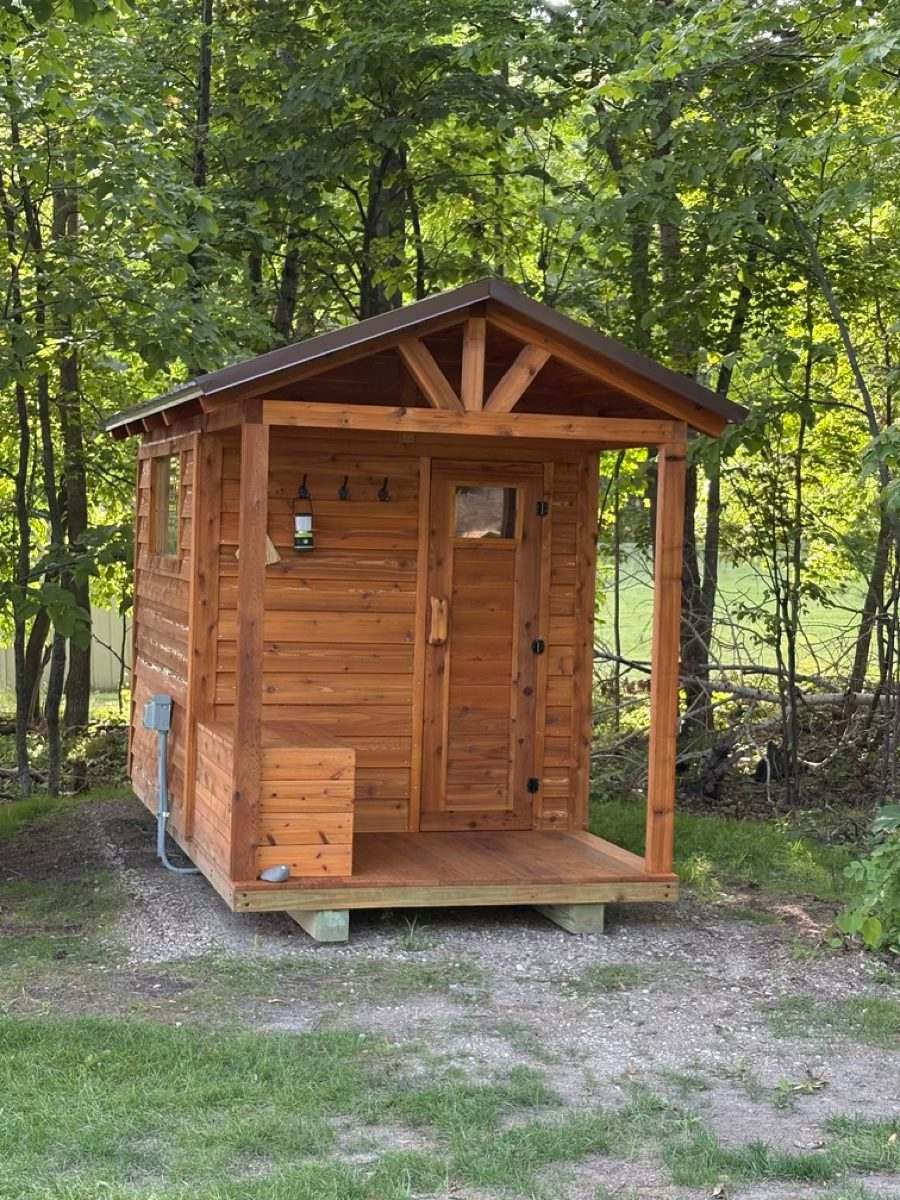 Front of a stained cedar sauna with a porch bench, lantern, towel hooks and a glass-panel door