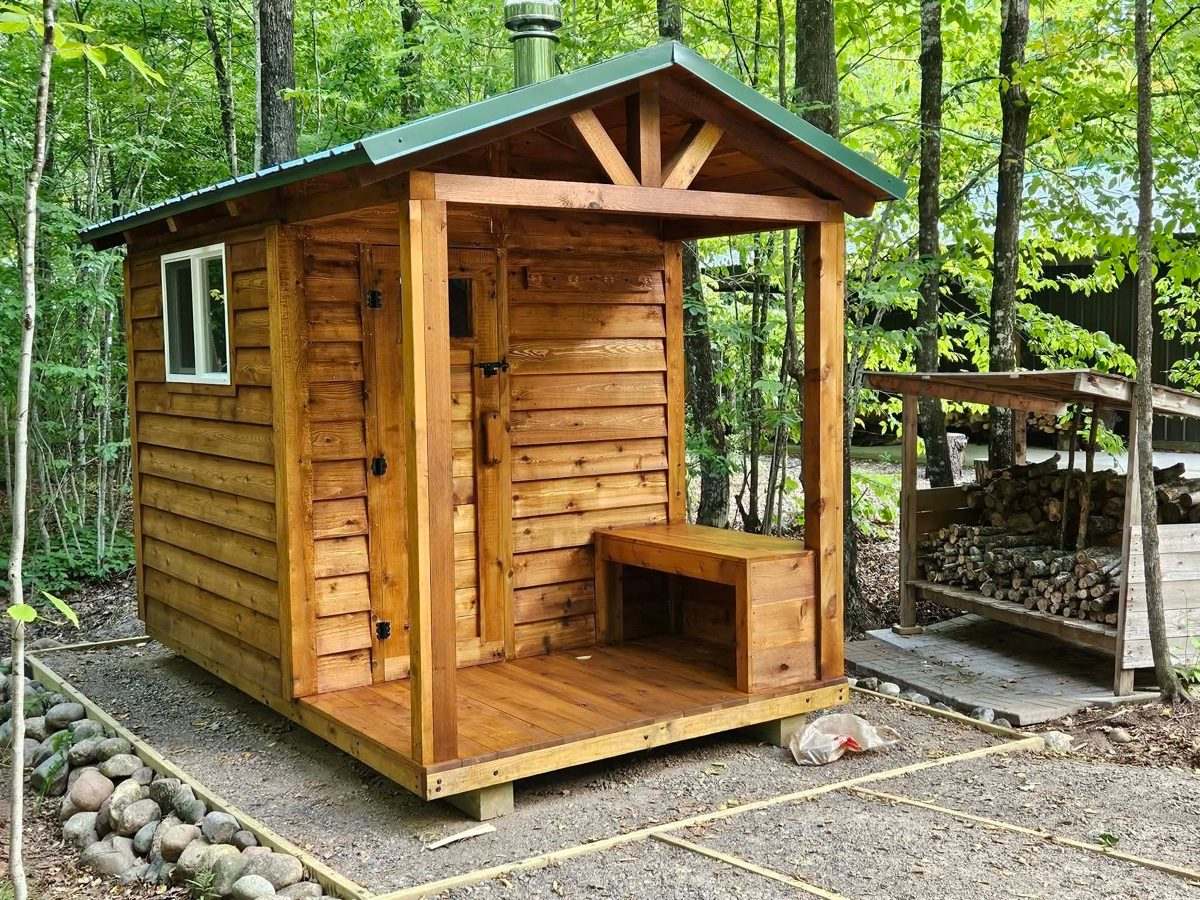Stained cedar outdoor sauna with a green metal roof, porch bench and a firewood shed beside it