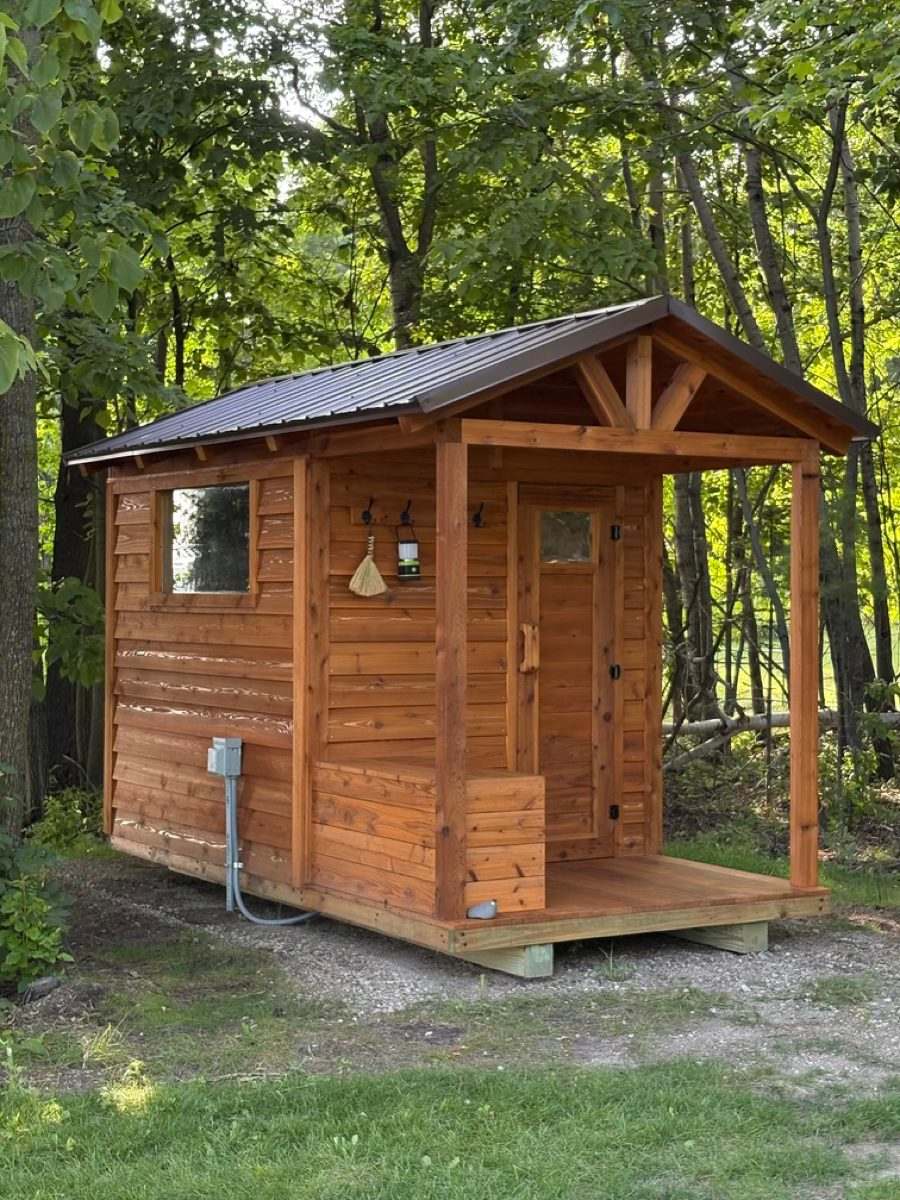 Stained cedar outdoor sauna with a brown metal roof, covered porch, bench and side window