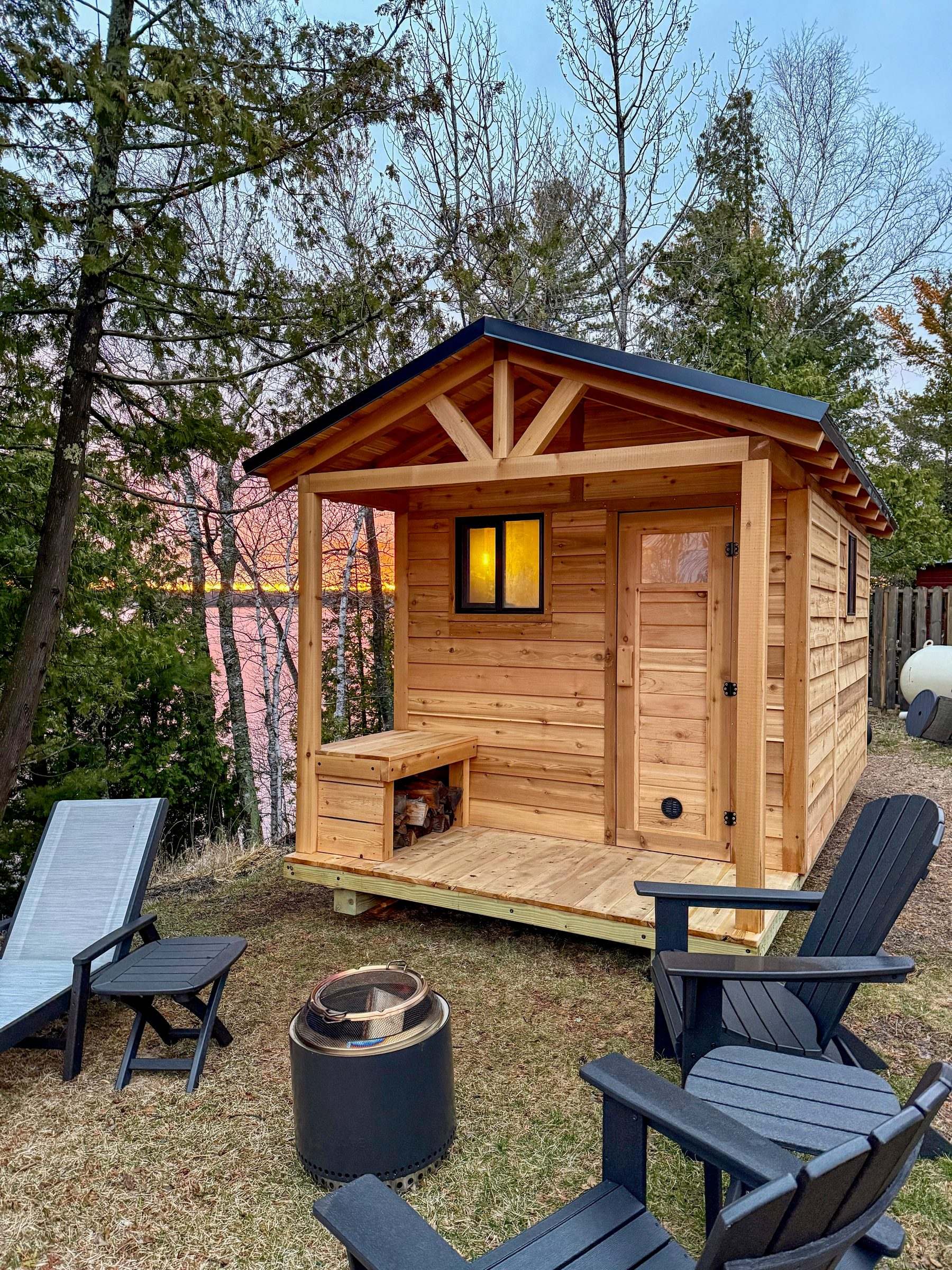 Cedar outdoor sauna with a glowing window at sunset beside a lake, with Adirondack chairs and a fire pit